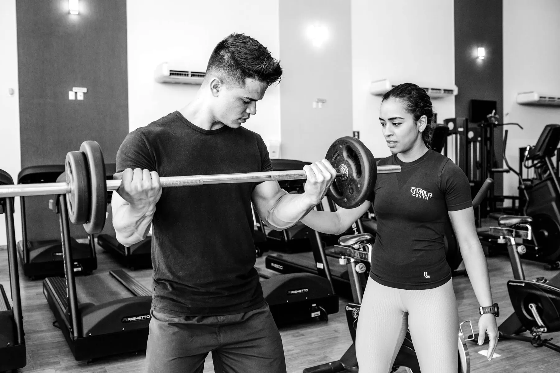 A personal trainer supporting a client lifting a barbell in the gym, black and white